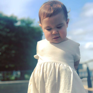 Mia, age 3, standing outside in a white dress on a sunny day.