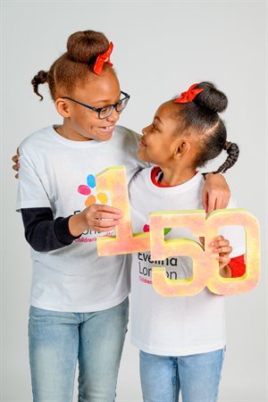 Tiana and her sister red bows smiling and looking at each other, holding a 150 sign.